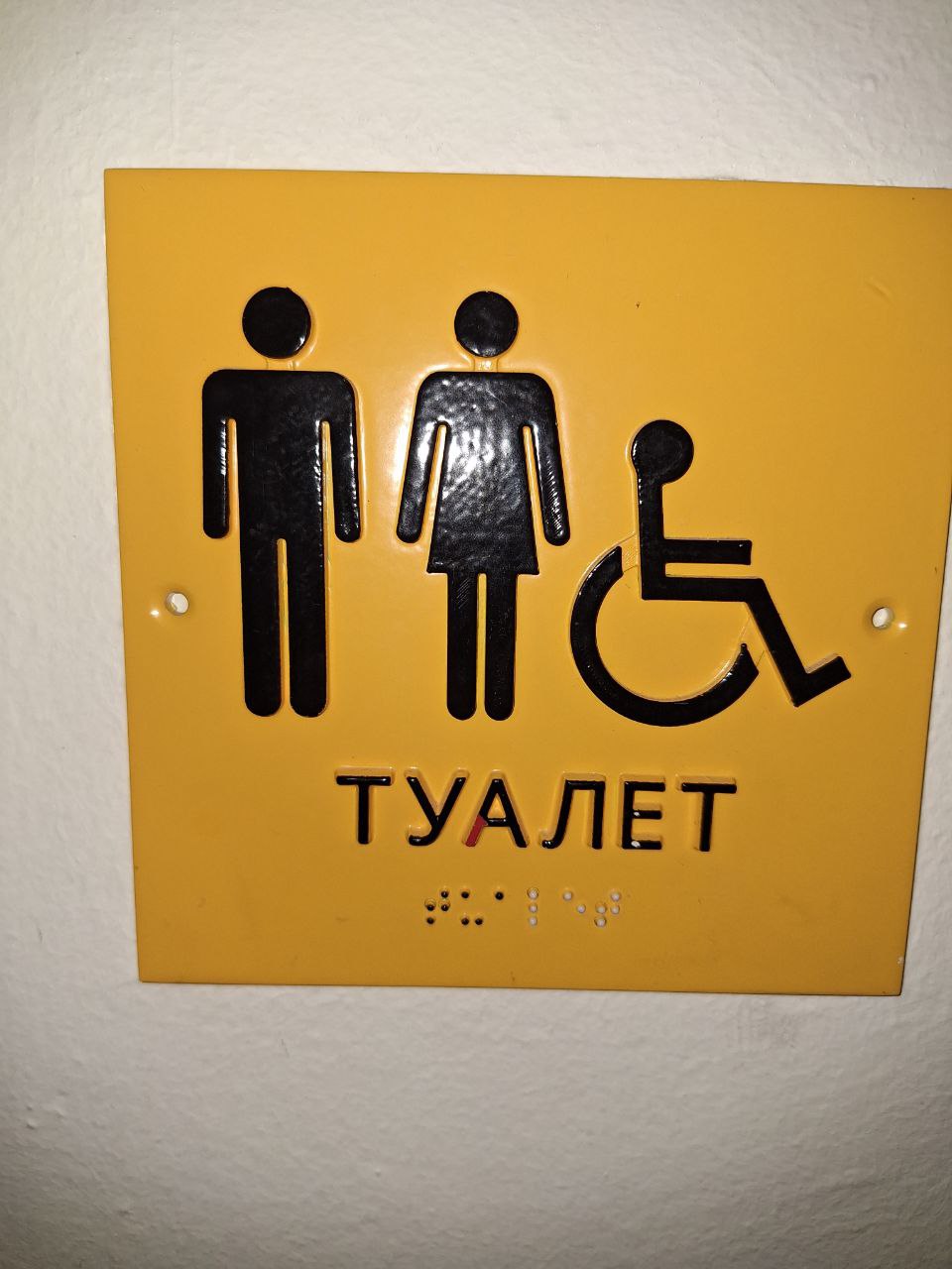 Жовта табличка із піктограмами чоловіка, жінки та символом доступності (людина на кріслі колісному), що вказує на ТУАЛЕТ, продубльовано шрифтом Брайля